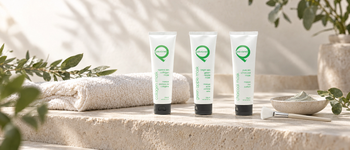 Three Pevonia facial mask tubes, Ageless Skin Collagen, Bright Skin Green Apple and Pure Skin Charcoal, arranged on a spa treatment bench with a facial brush, folded towel and greenery.