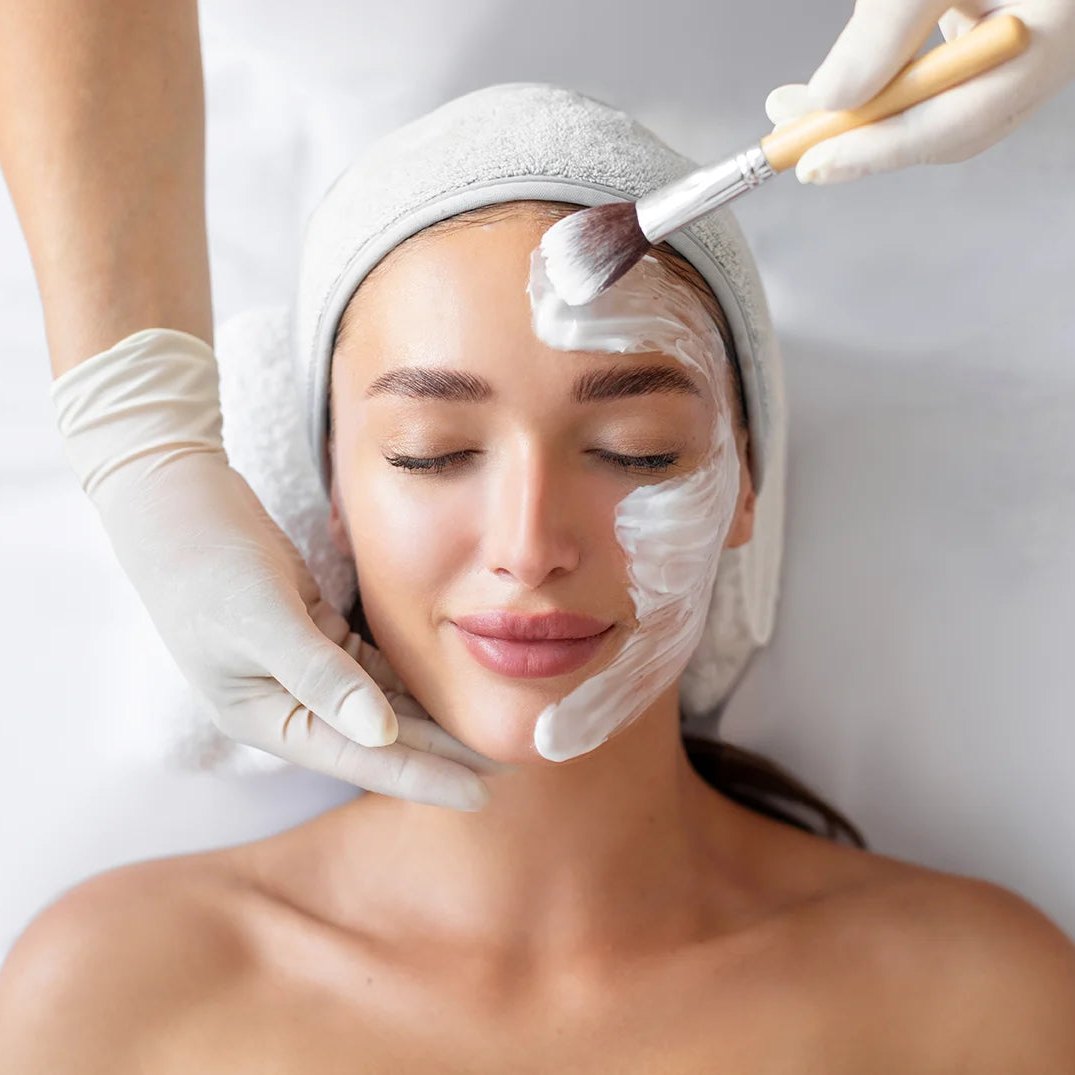 Woman receiving a facial treatment with a white towel on her head and a brush applying cream to her face.