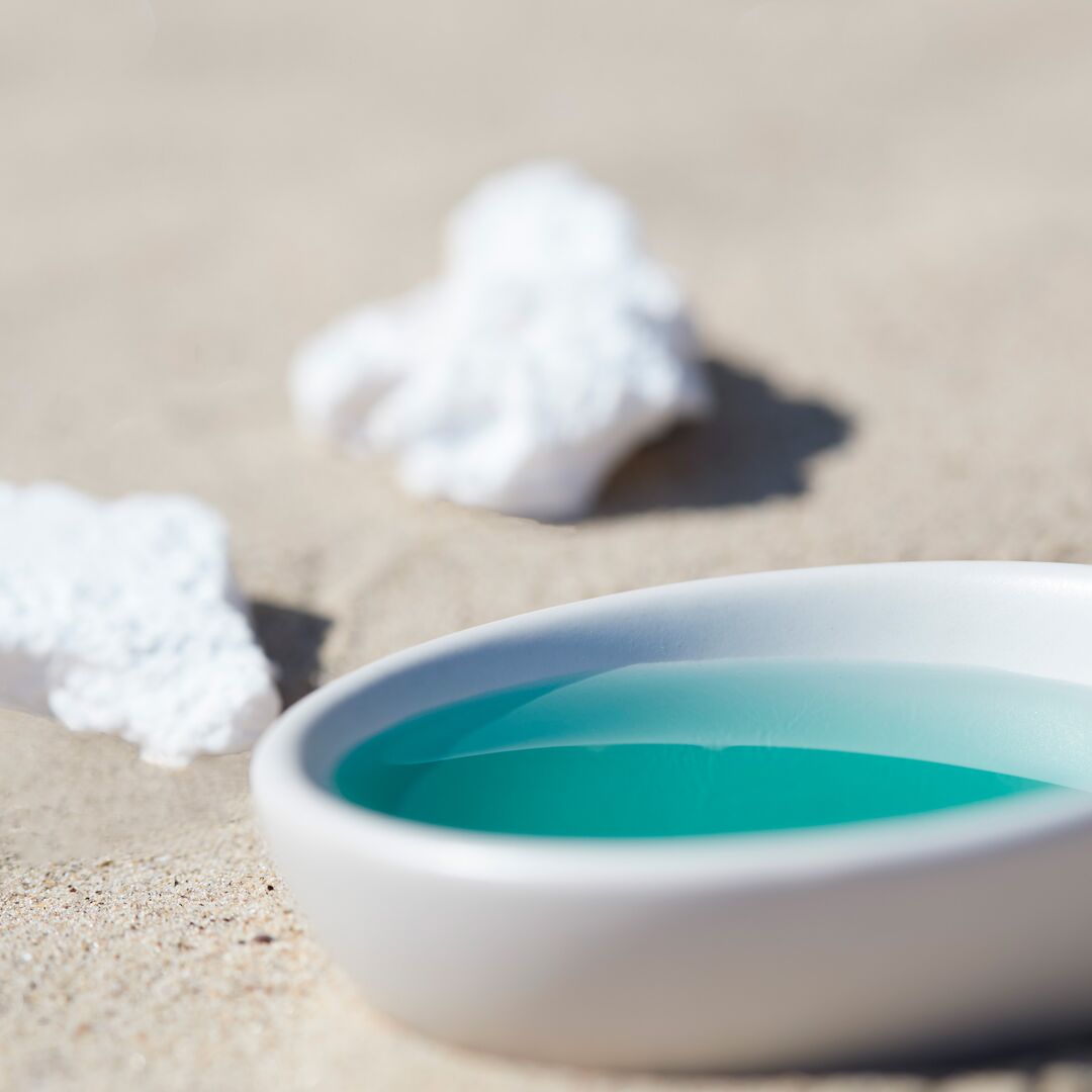 White dish with blue liquid on a sandy surface with shells.