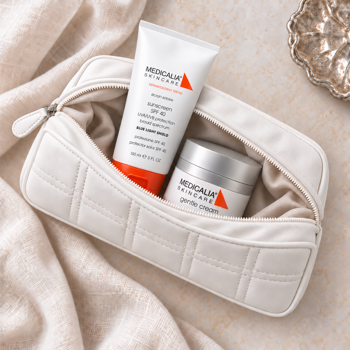 A white quilted cosmetic pouch with a zipper, shown open with two Medicalia Skincare products inside: a white and orange sunscreen tube and a white cream jar, arranged on draped beige fabric.