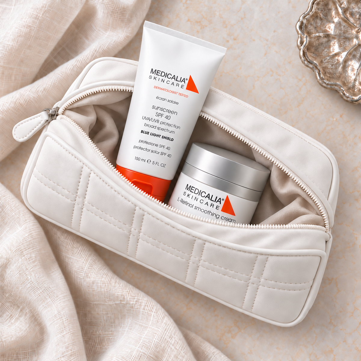 A quilted cream-colored cosmetic pouch with a zipper, shown open with two Medicalia skincare products inside: a white sunscreen tube with orange accents and a silver jar of face cream, arranged on a soft beige fabric surface.