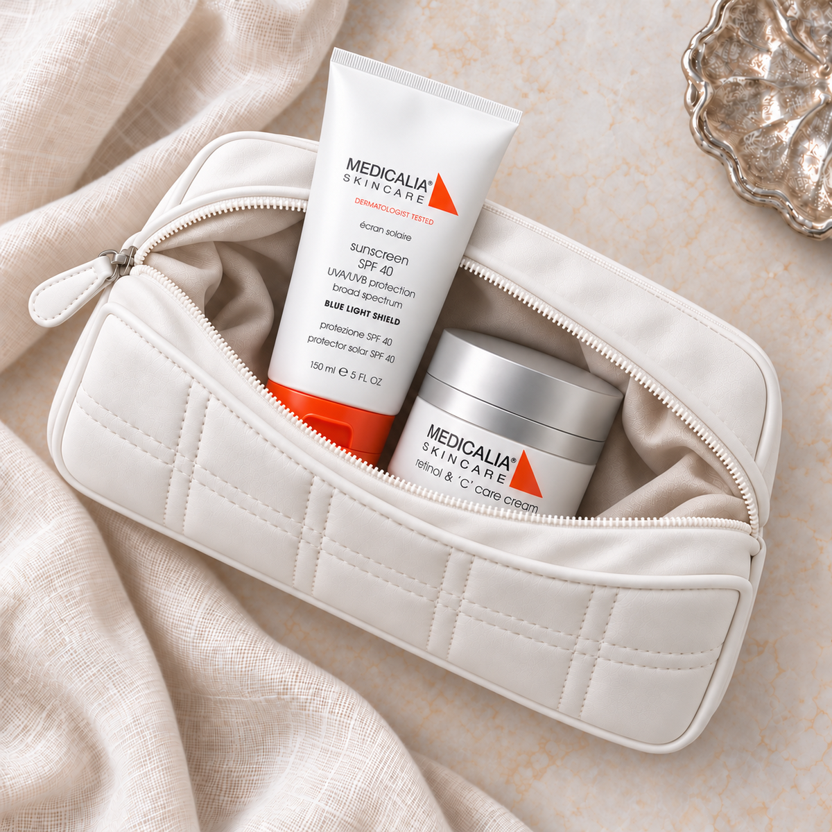 A Medicalia Retinol and C Care Cream & Sunscreen Gift Bag - white bag with a zipper, shown open and filled with Sunscreen  SPF 40 and a white jar labeled Retinol & C care cream. The pouch is styled on beige fabric with a decorative silver dish in the corner.