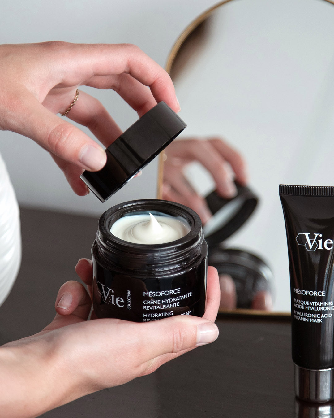 Person opening a jar of Vie Collection Mesoforce Hydrating Revitalizing Cream. Tube of Mesoforce Hyaluronic Acid Vitamin Mask visible.