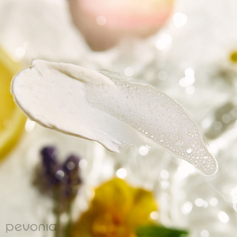 Pevonia Hyaluronic Acid Multi Active Hydra Cleanser indicating Texture and ingredients Close-up of a white cream smear with blurred flowers in the background, featuring the brand 'pevonia'.