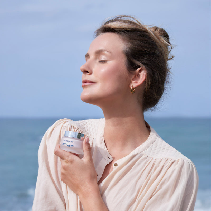Woman holding a skincare product with ocean and sky in the background