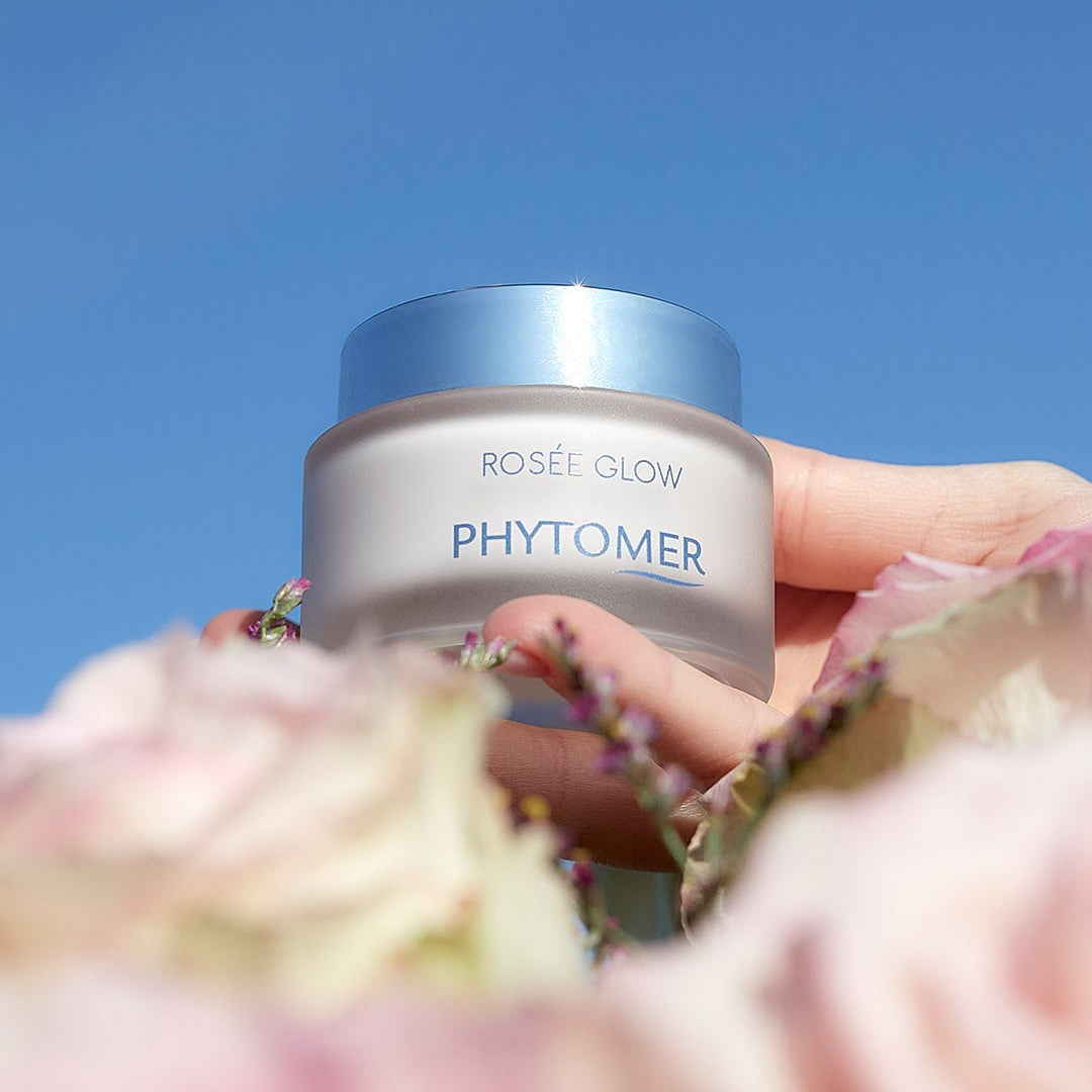 Phytomer Rosee Glow jar held against a blue sky with flowers in the foreground