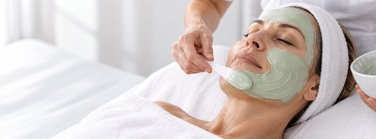 A woman lying down with a white towel wrapped around her head while a spa worker applies a light green facial mask with a spatula. A small bowl of mask powder is held on the right side.