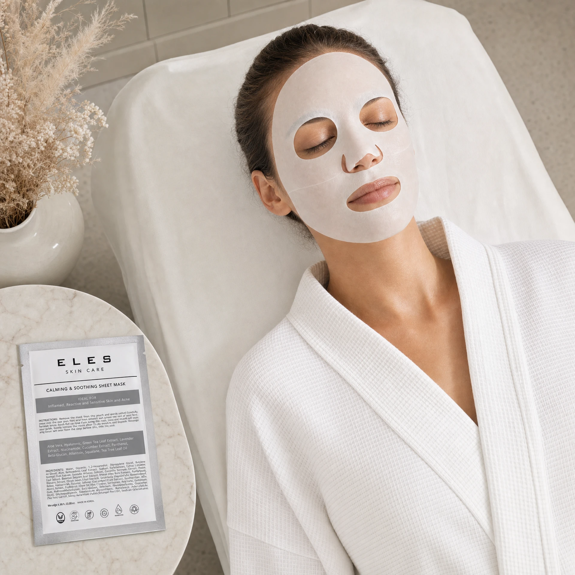 ELES Calming and Soothing Sheet Mask applied during a clinical skincare treatment, woman relaxing on treatment bed in white robe, silver foil sachet on marble side table, sensitive and reactive skin treatment