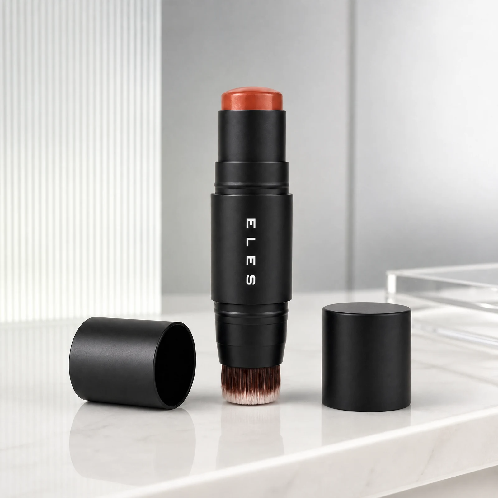 A black ELES cheek and lip cream with a coral-orange cosmetic bullet and visible brush bristles, standing upright on a white marble surface. The brand name 'ELES' is printed vertically on the tube.