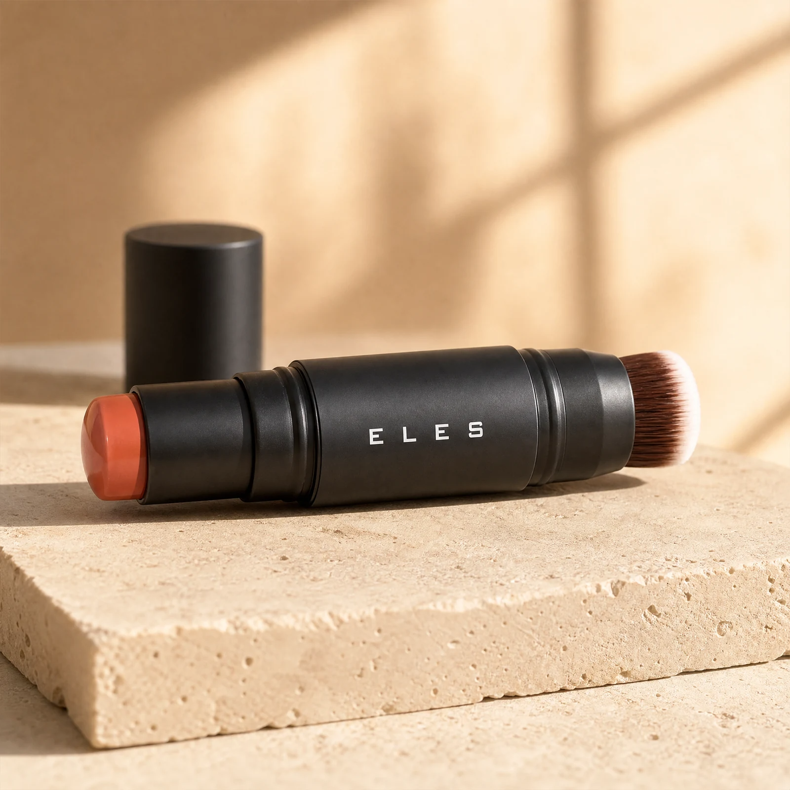 ELES cheek and lip cream, featuring a matte black body, a warm terracotta orange cosmetic product on one end, and dual-ended brown-and-white bristles on the other, displayed on a textured beige stone block with its cap standing behind it.
