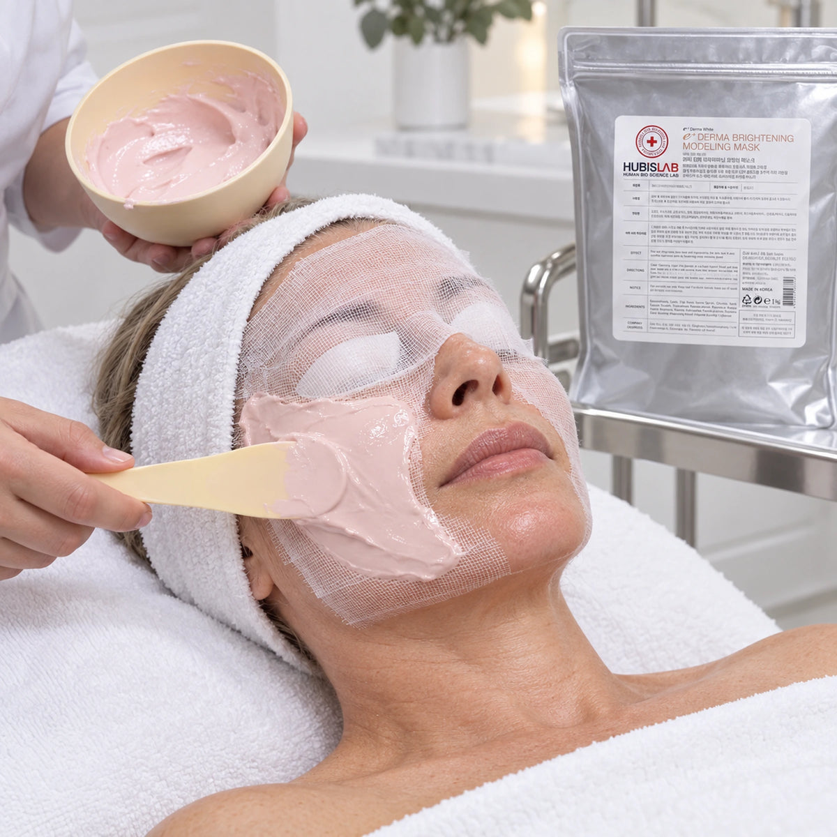 Therapist hands applying pale pink HUBISLAB Brightening Modeling Mask over gauze, with the 1 kg pouch beside the treatment bed.