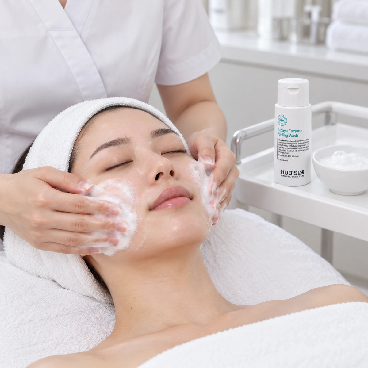 Therapist cleansing a client’s face with HUBISLAB Papaya Enzyme Peeling Wash, with the 70 g bottle on the trolley.