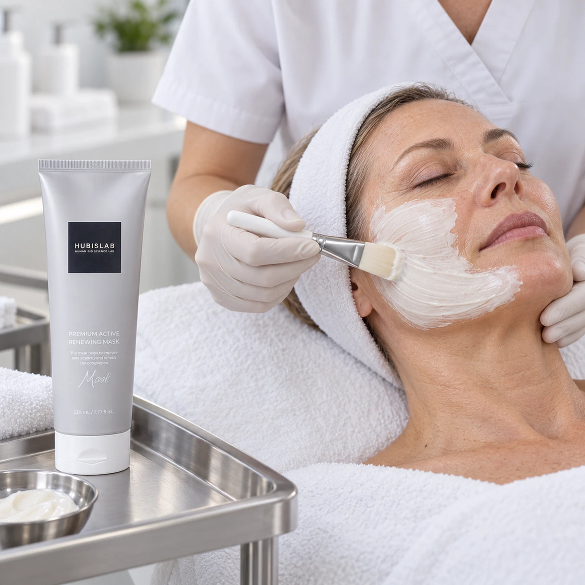 Therapist applying HUBISLAB Premium Active Renewing Mask during a facial, with the professional 230 ml tube on a tray.