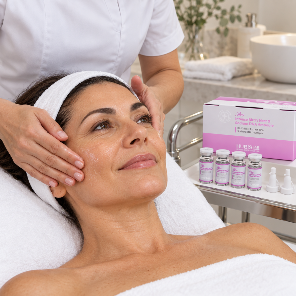 Therapist smoothing a clear serum over a client’s cheek during a professional facial, with the HUBISLAB R20 four-vial ampoule kit on the trolley.
