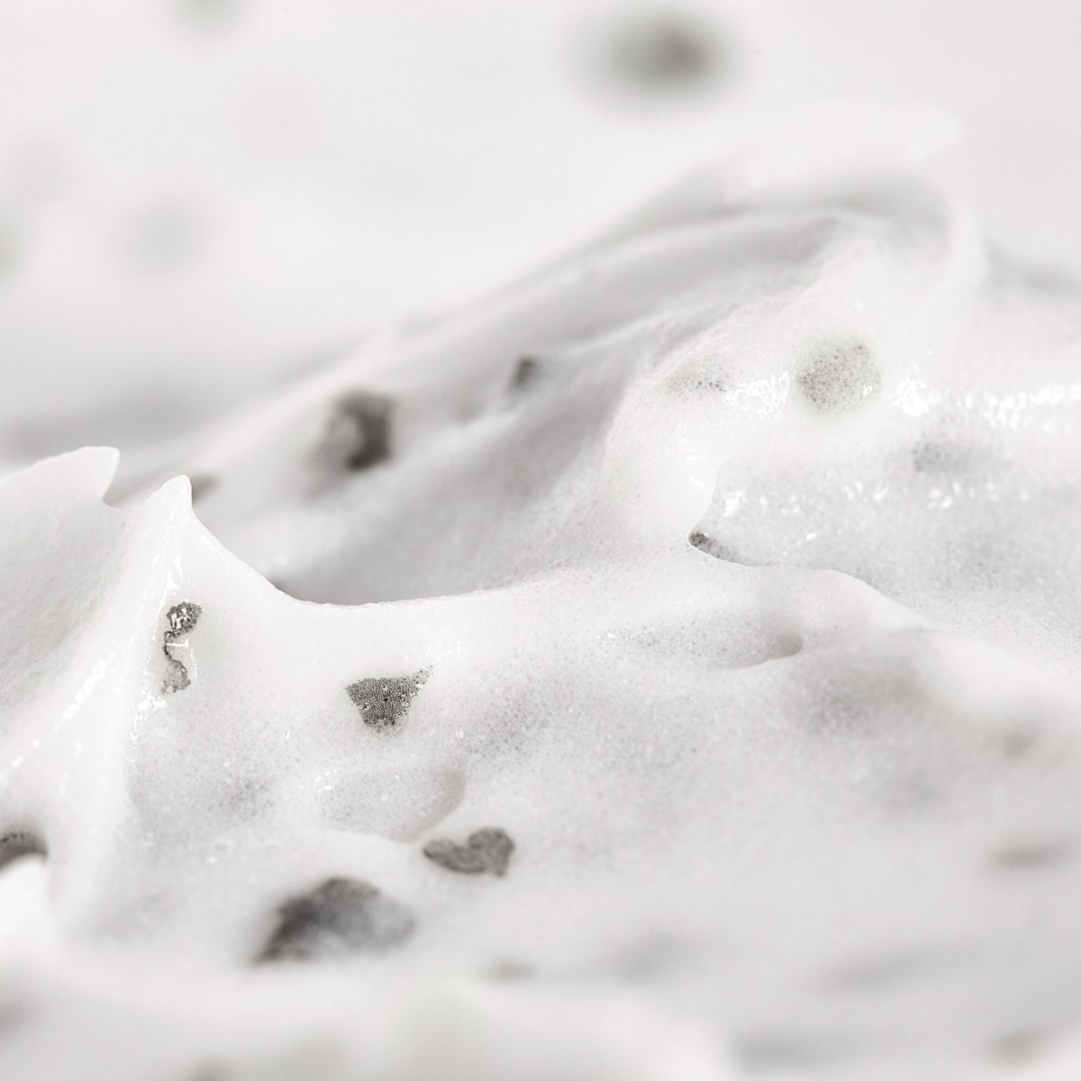 A close-up texture shot of a thick, creamy white cosmetic or food product, possibly a scrub, cream, or mousse, containing small dark gray and black flecks, with a soft-focus background.