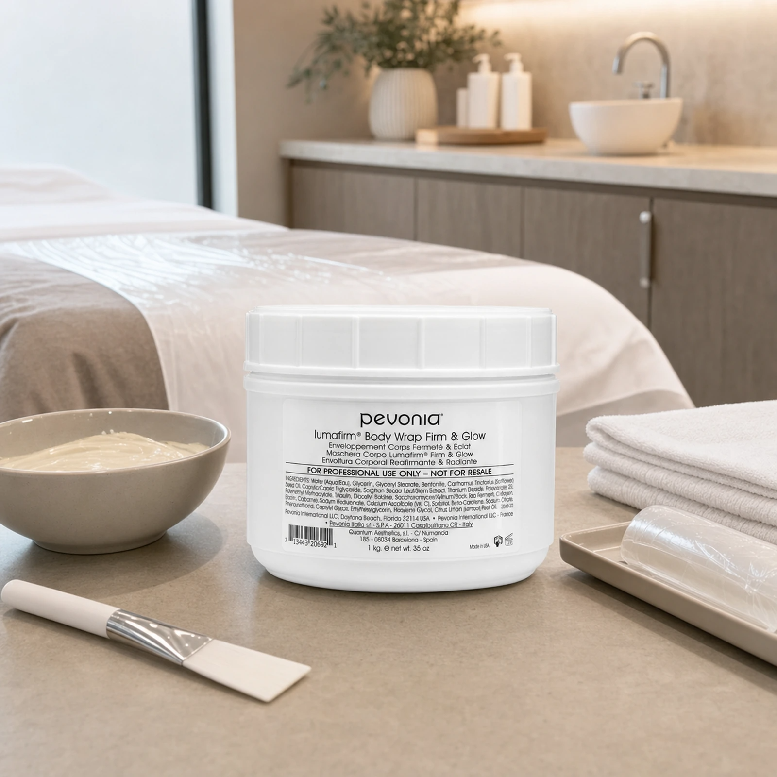 Pevonia Lumafirm Body Wrap Firm & Glow 1kg jar displayed in an upmarket clinic body treatment room with body wrap preparation accessories, folded towels and a therapist trolley.