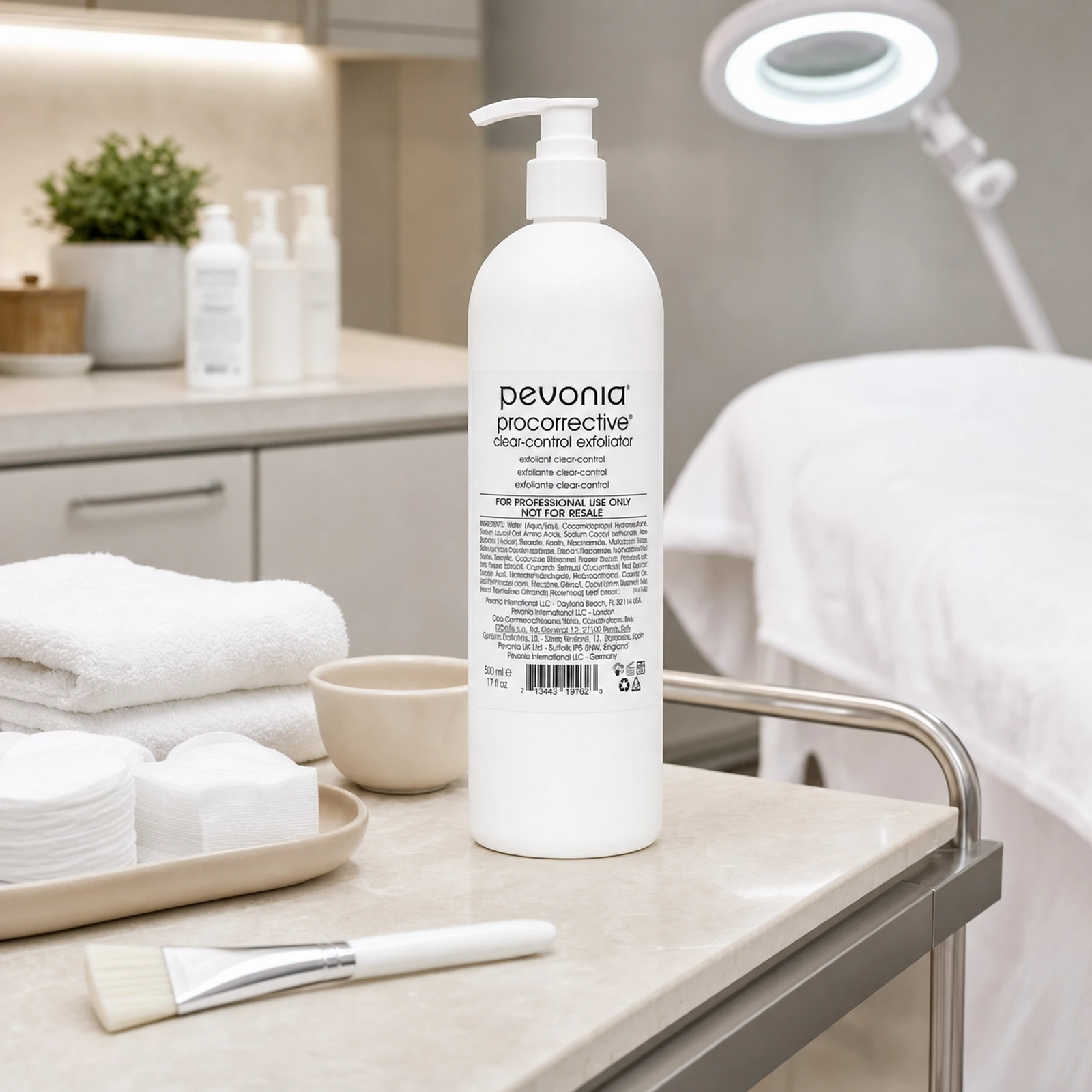 Pevonia ProCorrective Clear-Control Exfoliator 500g pump bottle displayed in an upmarket clinic facial treatment room with exfoliation preparation accessories, folded towels and a therapist trolley.