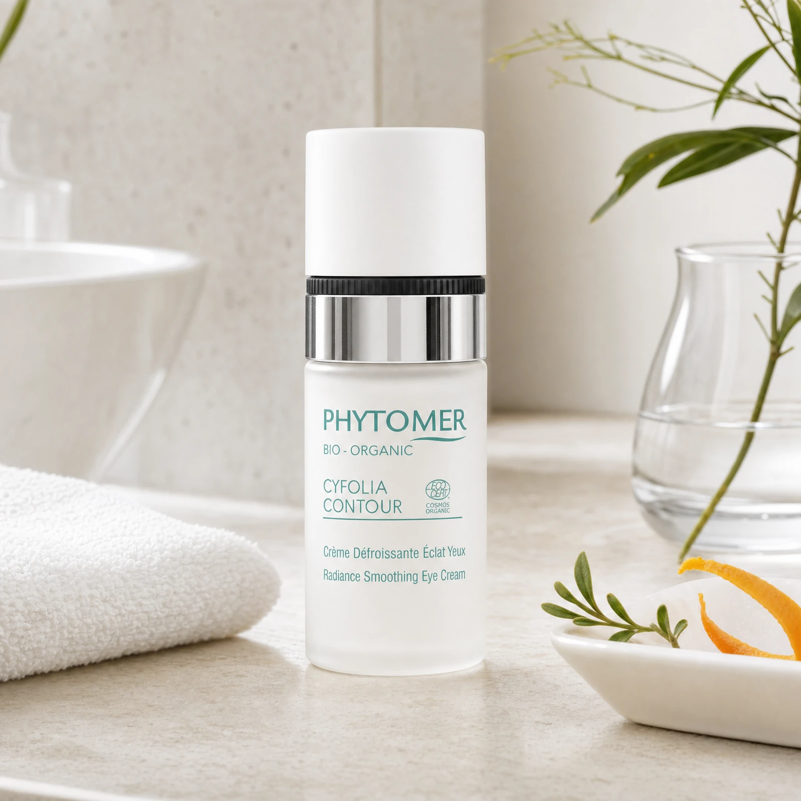 Phytomer Cyfolia Organic Radiance Smoothing Eye Cream 15ml on a marble bathroom counter with orange peel in ceramic dish, botanical in glass vase, rosemary sprig and white towels, premium at-home organic skincare setting