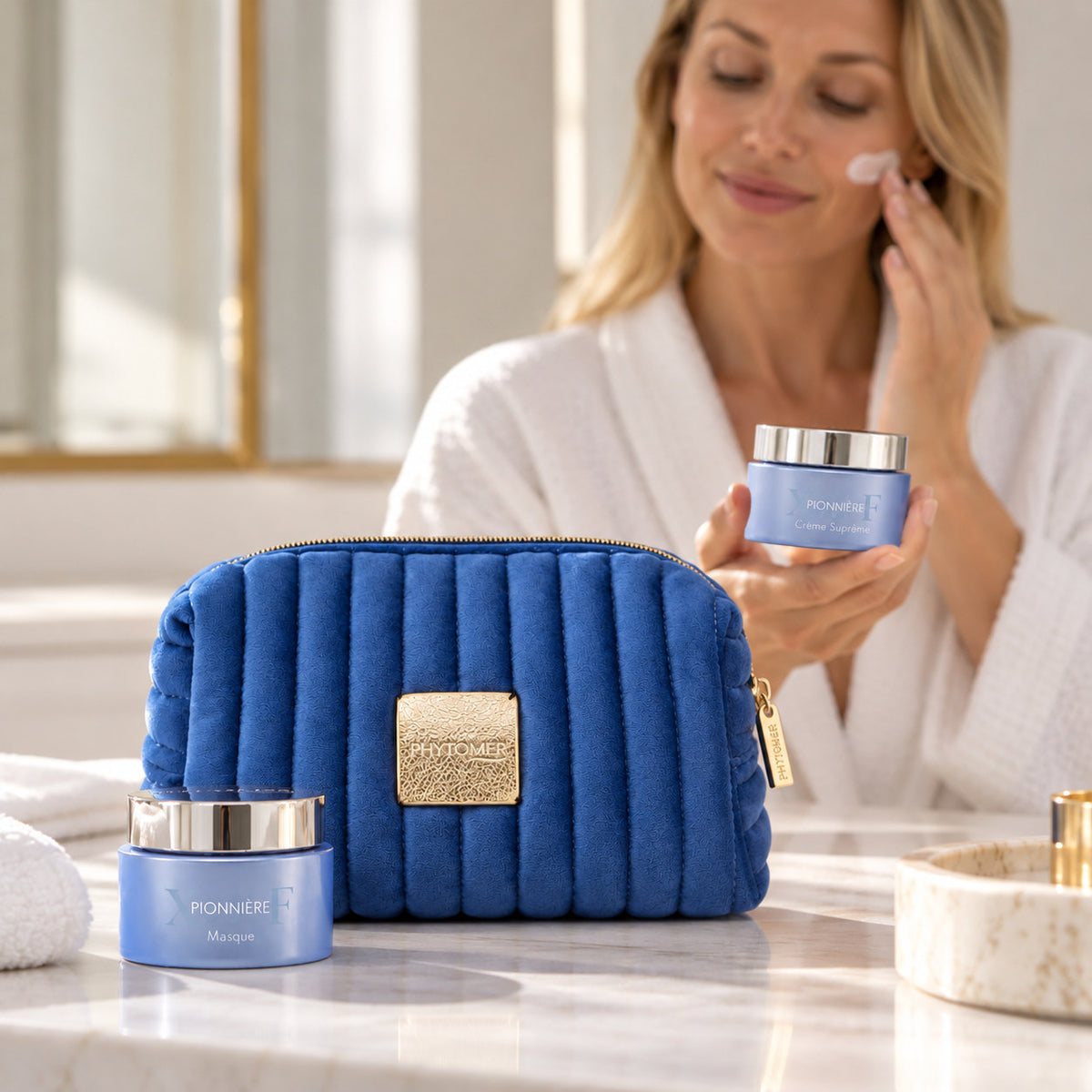 Woman applying PHYTOMER Pionnière XMF Supreme Cream beside the blue gift pouch and matching mask jar.