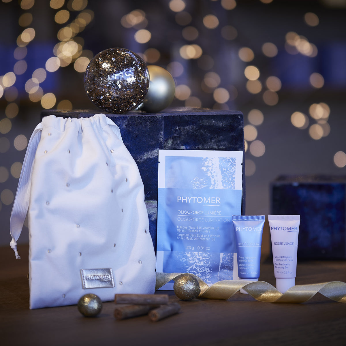 Phytomer Ritual Glow Gift Set with cleansing gel, moisturising cream, sheet mask and white pouch beside gold baubles and festive lights.