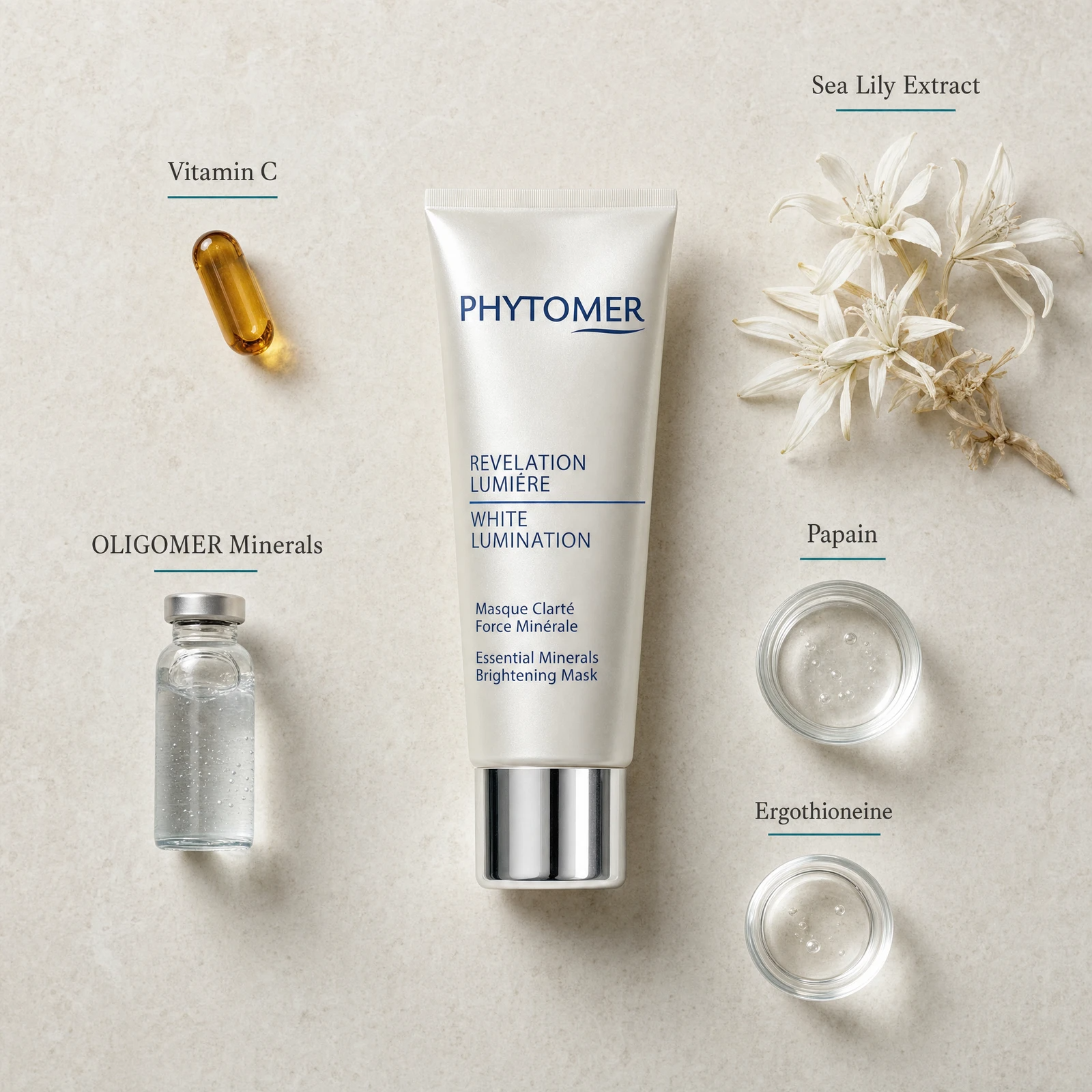 Phytomer Revelation Lumière White Lumination mask tube centered on a light beige textured background, surrounded by skincare ingredients including a Vitamin C capsule, oligomer minerals vial, sea lily extract flowers, papain, and ergothioneine dropper containers.