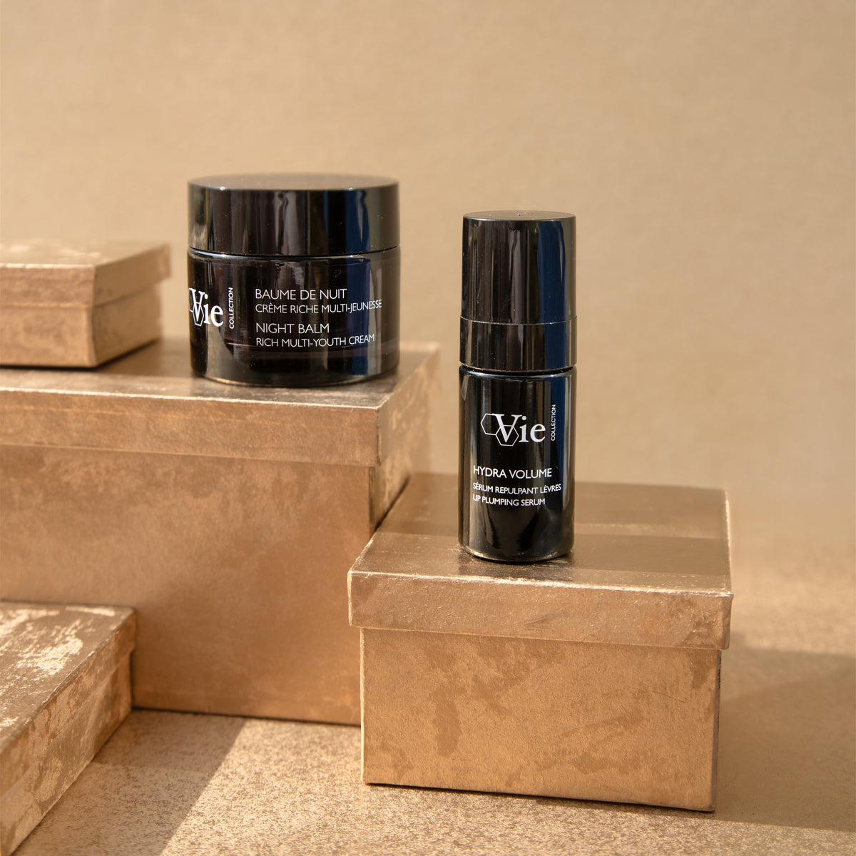 Vie Collection Night Balm and Hydra Volume Lip Plumping Serum arranged on stacked gold gift boxes against a gold backdrop.