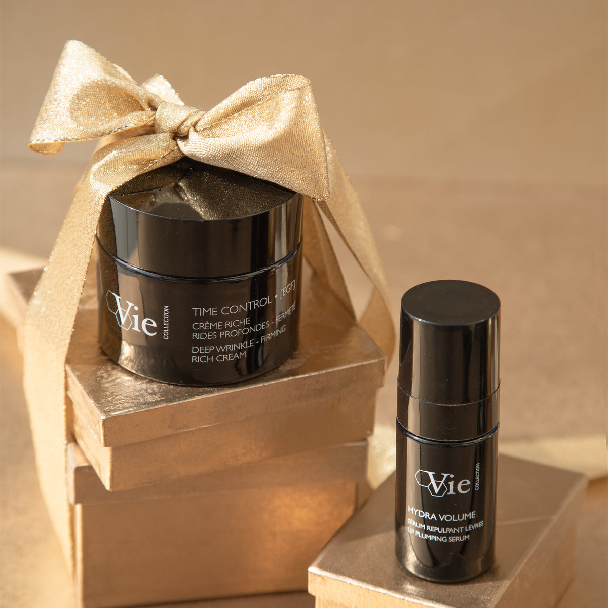 Vie Collection Time Control cream topped with a gold bow and Hydra Volume Lip Plumping Serum displayed on gold gift boxes.